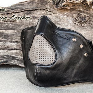 Puede incluir: Máscara facial de cuero negro con un panel de cuero gris perforado y el texto "Holler & Hood" impreso en el lateral.