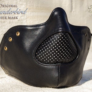 Pode incluir: Máscara de couro preta com o texto "The Original Thunderbird Leather Mask". A máscara tem uma área para os olhos perfurada e rebites dourados. A marca "Holler & Hood" é visível no canto superior direito.