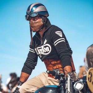 Könnte beinhalten: Ein Mann mit einem blau-weiß-roten Helm mit einer braunen Leder-Gesichtsmaske, einem schwarz-weiß gestreiften Pullover mit dem Wort "Triton" darauf und einem braunen Ledergürtel. Er sitzt auf einem Motorrad.