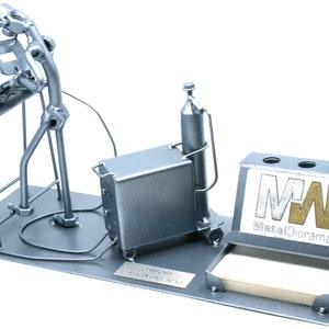 May include: A silver metal desk organizer featuring a figurine of a welder working on a project. The organizer includes a pen holder, a note pad holder, and a small metal box. The figurine is made of nuts, bolts, and other metal pieces. The organizer has a metal plate with the text "MetalDorama" and the initials "MW".