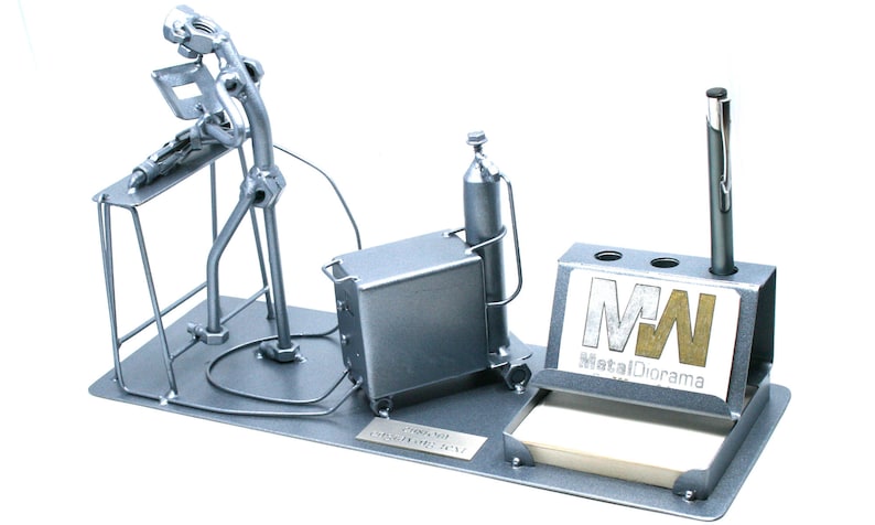 May include: A silver metal desk organizer featuring a figurine of a welder working on a project. The organizer includes a pen holder, a note pad holder, and a small metal box. The figurine is made of nuts, bolts, and other metal pieces. The organizer has a metal plate with the text "MetalDorama" and the initials "MW".