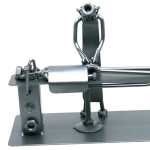 May include: A silver-colored metal sculpture crafted from nuts, bolts, and metal scraps, depicting a figure standing over another figure lying on a flat rectangular platform. The entire assembly is welded together and mounted on a flat base.
