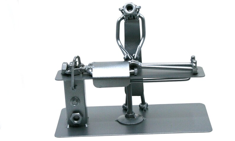 May include: A silver-colored metal sculpture crafted from nuts, bolts, and metal scraps, depicting a figure standing over another figure lying on a flat rectangular platform. The entire assembly is welded together and mounted on a flat base.