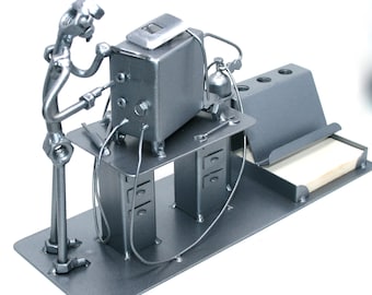 May include: A silver metal sculpture depicting a person working on a machine at a desk. The sculpture includes a notepad holder and a small tank. The figure is made of nuts and bolts.