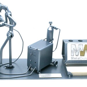 May include: A metal figurine of a welder working on a metal table with a welding torch, a welding machine, and a pen holder with the logo "MetalDiorama" on it.