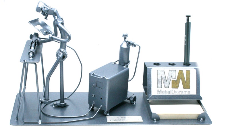 May include: A metal figurine of a welder working on a metal table with a welding torch, a welding machine, and a pen holder with the logo "MetalDiorama" on it.