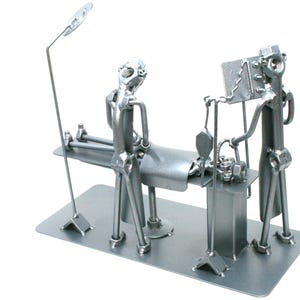 May include: A silver-colored metal sculpture depicting a surgical scene. The piece features two figures standing over a patient lying on an operating table. The sculpture includes a tall surgical lamp, an IV pole, and a monitor, all crafted from hardware components.