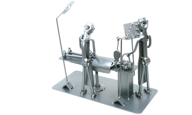 May include: A silver-colored metal sculpture depicting a surgical scene. The piece features two figures standing over a patient lying on an operating table. The sculpture includes a tall surgical lamp, an IV pole, and a monitor, all crafted from hardware components.