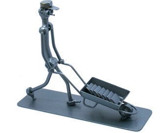 May include: A metal sculpture of a worker pushing a wheelbarrow. The worker is made of metal rods and bolts, wearing a cap. The wheelbarrow is filled with rectangular blocks. The sculpture is on a rectangular base.