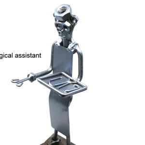 May include: A silver-colored metal figurine of a surgical assistant constructed from hardware components including nuts and bolts. The figure holds a small rectangular tray with surgical tools and has the text surgical assistant printed nearby.