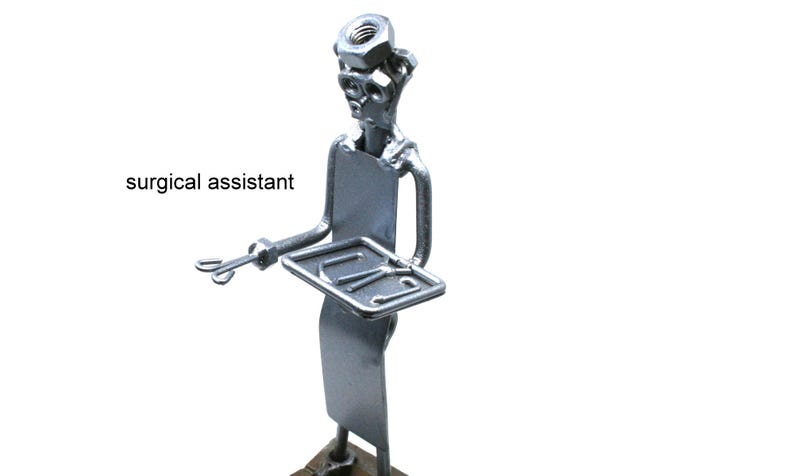 May include: A silver-colored metal figurine of a surgical assistant constructed from hardware components including nuts and bolts. The figure holds a small rectangular tray with surgical tools and has the text surgical assistant printed nearby.