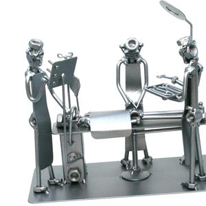 May include: A silver-toned metal sculpture depicting a surgical scene with three figures constructed from nuts, bolts, and metal scraps. The figures stand around a central operating table, with one figure holding a tray and another near a lamp.