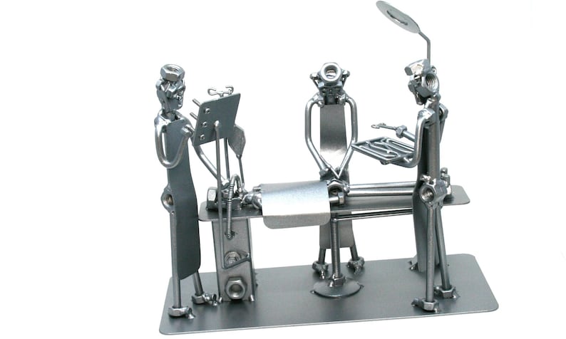 May include: A silver-toned metal sculpture depicting a surgical scene with three figures constructed from nuts, bolts, and metal scraps. The figures stand around a central operating table, with one figure holding a tray and another near a lamp.