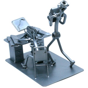 Puede incluir: Una escultura de metal de dos figuras, una sentada en un escritorio con un monitor de computadora y la otra corriendo. Las figuras están hechas de tuercas, tornillos y otras piezas de metal.