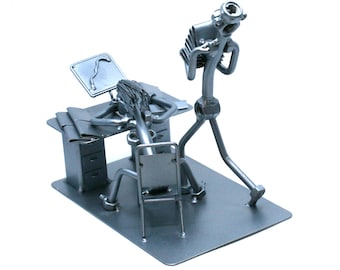 May include: A metal sculpture of two figures, one sitting at a desk with a computer monitor and the other running away. The figures are made of nuts, bolts, and other metal pieces.