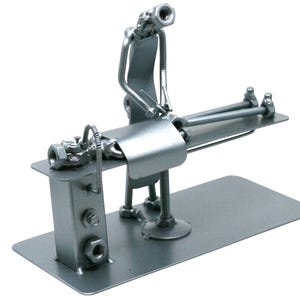 May include: A silver-colored metal sculpture crafted from industrial hardware components, including nuts, bolts, and metal plates. The figure depicts a person standing beside a table, appearing to perform a medical procedure on a reclining figure.