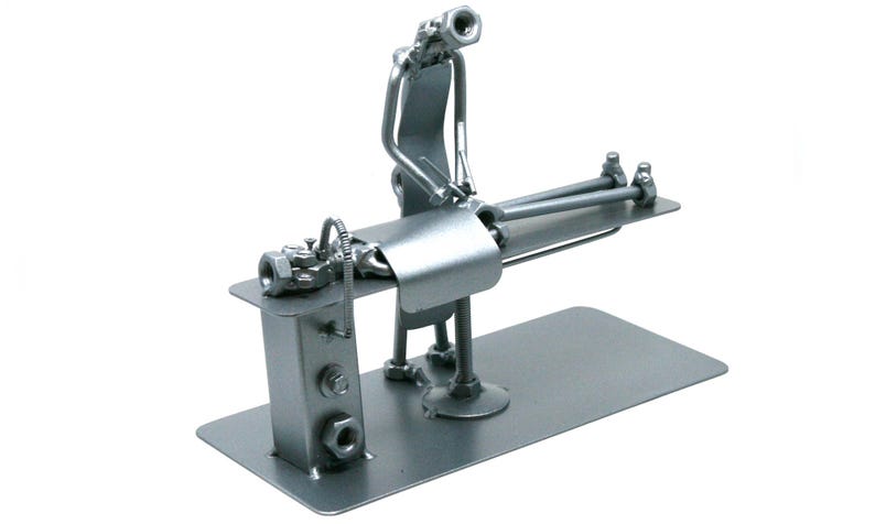 May include: A silver-colored metal sculpture crafted from industrial hardware components, including nuts, bolts, and metal plates. The figure depicts a person standing beside a table, appearing to perform a medical procedure on a reclining figure.