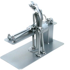 May include: A silver-colored metal sculpture constructed from various hardware components, including nuts, bolts, and rods. The figure is depicted in a seated position on a flat rectangular base, with legs extended forward and arms resting on the surface.