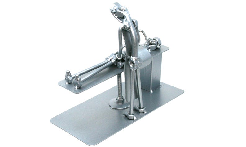 May include: A silver-colored metal sculpture constructed from various hardware components, including nuts, bolts, and rods. The figure is depicted in a seated position on a flat rectangular base, with legs extended forward and arms resting on the surface.
