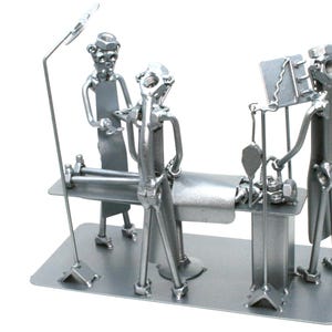 May include: A silver-toned metal sculpture depicting a surgical scene with three figures made from nuts, bolts, and metal scraps. The figures are positioned around an operating table, with one figure standing by a tall lamp and another by a monitor.