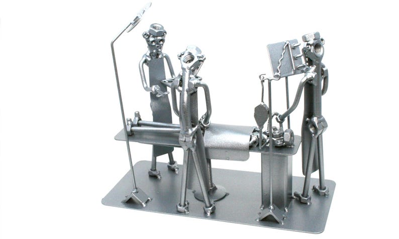 May include: A silver-toned metal sculpture depicting a surgical scene with three figures made from nuts, bolts, and metal scraps. The figures are positioned around an operating table, with one figure standing by a tall lamp and another by a monitor.