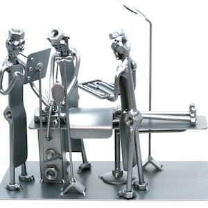 May include: A silver-toned metal sculpture depicting a medical scene with three figures constructed from nuts, bolts, and metal scraps. The figures are positioned around an operating table with an IV stand and a surgical light fixture overhead.