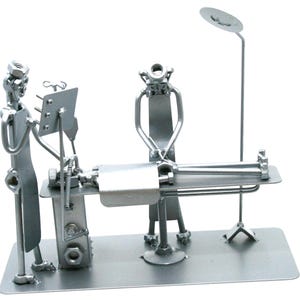 May include: A silver-colored metal sculpture depicting a medical scene with two figures standing over a patient on an operating table. The figures are constructed from various hardware components like nuts, bolts, and metal rods, featuring a tall lamp.