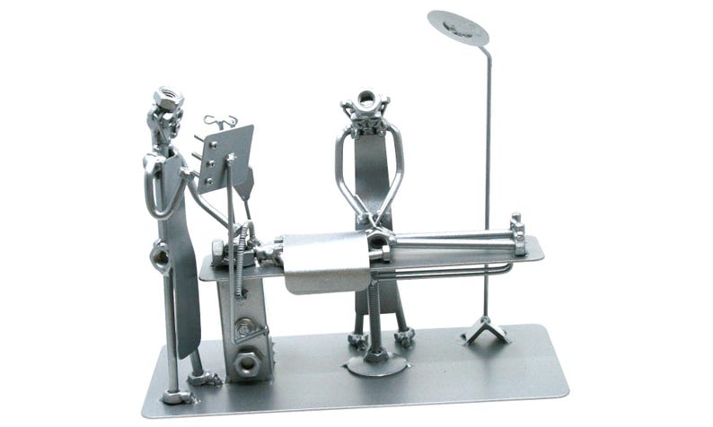 May include: A silver-colored metal sculpture depicting a medical scene with two figures standing over a patient on an operating table. The figures are constructed from various hardware components like nuts, bolts, and metal rods, featuring a tall lamp.