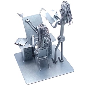 May include: A silver metal sculpture of two figures, one sitting at a desk with a lamp and the other standing and holding a piece of paper. The figures are made of wire and have long, flowing hair.