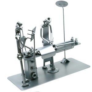 May include: A silver-colored metal sculpture depicting a medical examination scene. The piece is constructed from various hardware components including nuts, bolts, and metal rods, featuring a patient lying on a table with a medical professional.