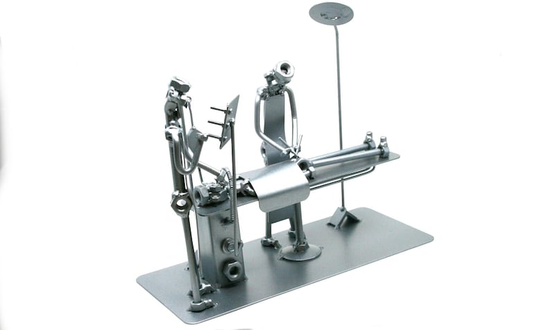 May include: A silver-colored metal sculpture depicting a medical examination scene. The piece is constructed from various hardware components including nuts, bolts, and metal rods, featuring a patient lying on a table with a medical professional.