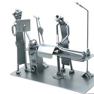May include: A silver-colored metal sculpture depicting a medical scene with two figures standing over a patient lying on an operating table. The figures are constructed from nuts, bolts, and metal rods, featuring an IV pole and a monitor screen.