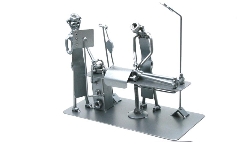 May include: A silver-colored metal sculpture depicting a medical scene with two figures standing over a patient lying on an operating table. The figures are constructed from nuts, bolts, and metal rods, featuring an IV pole and a monitor screen.