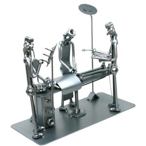 May include: A silver-toned metal sculpture depicting a medical surgery scene. Three figures constructed from nuts, bolts, and metal rods surround a central operating table. One figure stands at the head, another at the side, and one at the foot.