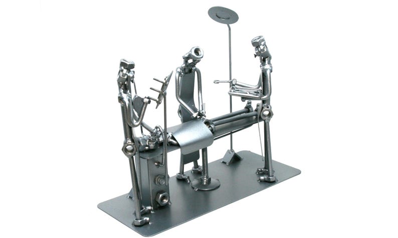 May include: A silver-toned metal sculpture depicting a medical surgery scene. Three figures constructed from nuts, bolts, and metal rods surround a central operating table. One figure stands at the head, another at the side, and one at the foot.