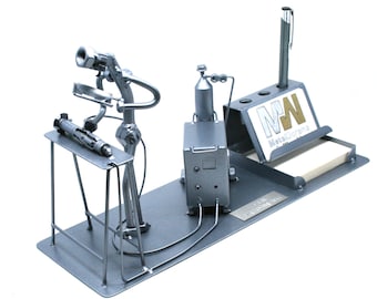 May include: A silver metal desk organizer shaped like a welding station. The organizer includes a pen holder, a business card holder, and a note pad holder. The organizer is made by MetalDreams.