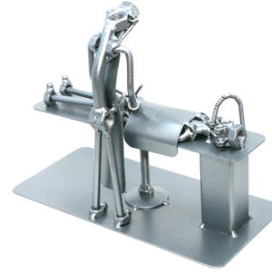 May include: A silver-colored metal sculpture constructed from industrial hardware components including nuts, bolts, and rods. The figure is positioned over a flat rectangular base, appearing to perform a task on a raised platform with a curved metal sheet.
