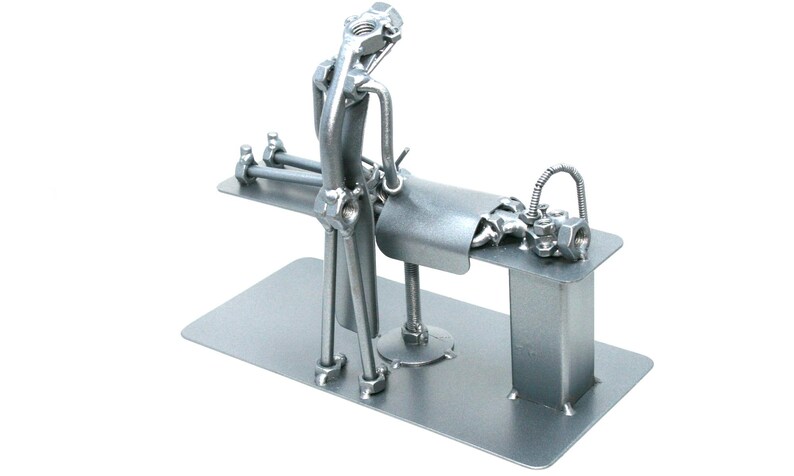 May include: A silver-colored metal sculpture constructed from industrial hardware components including nuts, bolts, and rods. The figure is positioned over a flat rectangular base, appearing to perform a task on a raised platform with a curved metal sheet.