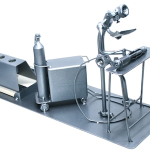 May include: A silver metal desk organizer shaped like a welder working on a table. The organizer has a pen holder, a small box, and a cylinder.
