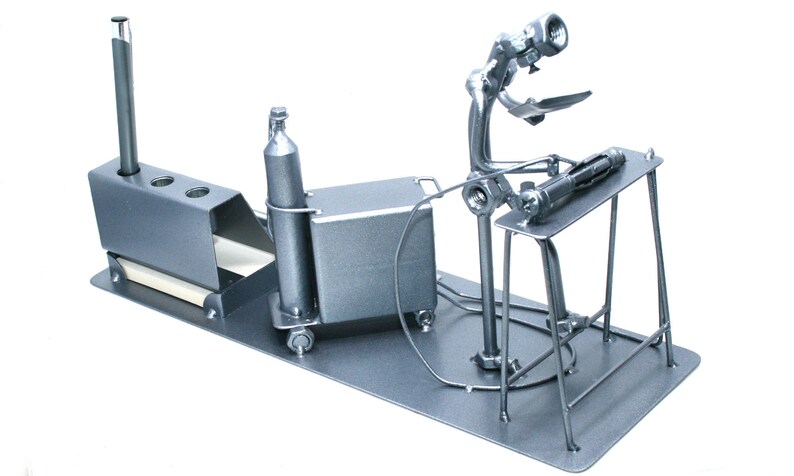 May include: A silver metal desk organizer shaped like a welder working on a table. The organizer has a pen holder, a small box, and a cylinder.