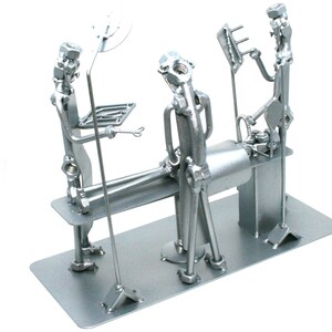 May include: A silver-colored metal sculpture depicting a surgical scene constructed from hardware components like nuts, bolts, and metal rods. The piece features figures representing medical staff around an operating table on a rectangular base.