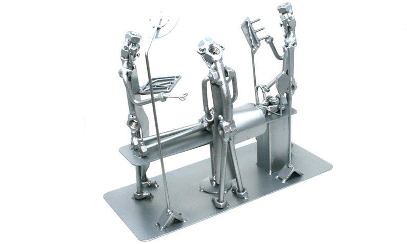 May include: A silver-colored metal sculpture depicting a surgical scene constructed from hardware components like nuts, bolts, and metal rods. The piece features figures representing medical staff around an operating table on a rectangular base.