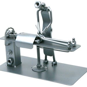 May include: A silver-colored metal sculpture crafted from hardware components like nuts, bolts, and metal strips. The piece depicts a figure standing over another figure lying on a flat rectangular platform, resembling a medical or massage table.