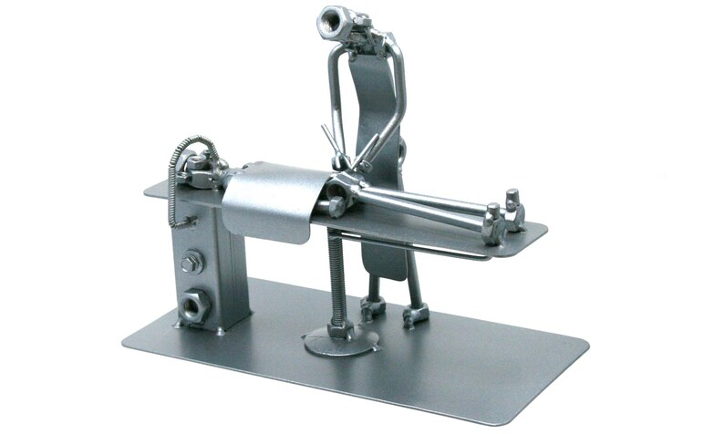 May include: A silver-colored metal sculpture crafted from hardware components like nuts, bolts, and metal strips. The piece depicts a figure standing over another figure lying on a flat rectangular platform, resembling a medical or massage table.