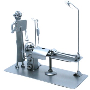 Op de afbeelding: Een metalen sculptuur van een dokter die een patiënt op een operatietafel onderzoekt. De sculptuur is gemaakt van moeren, bouten en andere metalen onderdelen. De dokter draagt een hoed en houdt een klembord vast. De patiënt ligt met uitgestrekte armen op de tafel. De sculptuur is een eigenzinnige en humoristische weergave van een medische scène.