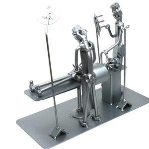 May include: A silver-toned metal sculpture depicting a medical scene with two figures constructed from nuts, bolts, and metal hardware. One figure stands over a patient lying on a table, while the other figure holds a medical instrument.