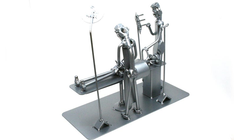 May include: A silver-toned metal sculpture depicting a medical scene with two figures constructed from nuts, bolts, and metal hardware. One figure stands over a patient lying on a table, while the other figure holds a medical instrument.