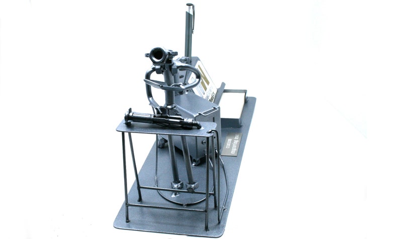 May include: A metal sculpture of a person sitting at a desk with a gun. The sculpture is made of recycled metal and is painted in a silver color. The sculpture is on a black base with the words "Art from Scrap" written on it.