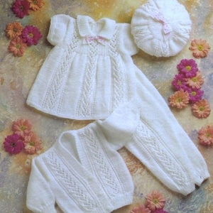 May include: A white knitted baby outfit set including a dress, cardigan, pants, and a beret. The set features a delicate lace pattern and a pink bow on the dress.