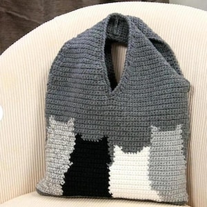 May include: A gray crochet tote bag featuring a cat design in shades of gray, black, and white. The bag has a V-neck opening and a wide strap for carrying. The handmade bag is sitting on a cream-colored corduroy chair.
